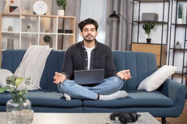 Hoş bir Arap genç Hintli adam, oturma odasındaki kanepede dizüstü bilgisayarla poz veriyor, ve meditasyon yapıyor, gözleri kapalı gülümsüyor, parmaklarını Mudra 'da kavuşturuyor.