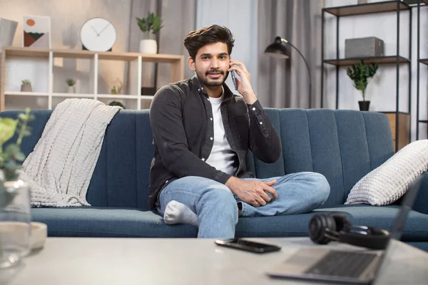 Smiling Indian man, sitting relaxed on comfortable sofa at living room, and talking on phone with call center operator or friend, making food order. People, domestic life and gadgets
