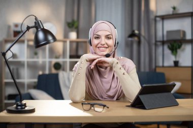 Oldukça tatmin olmuş tesettürlü, tesettürlü, tesettürlü, modern dekore edilmiş oturma odasındaki masada oturan ve kameraya bakan Arap genç bayan. Çevrimiçi iş, vlog veya webinar kavramı