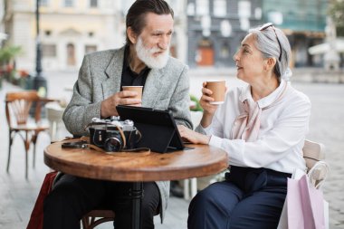 Kafe terasında kahve ve tabletle dinlenen yaşlı bir çift.