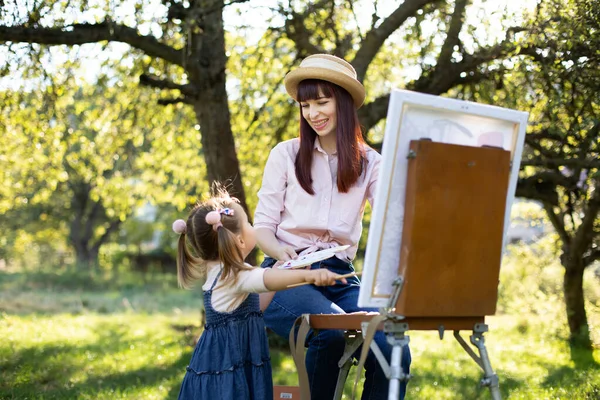 Recreation and family summer games in the open air. Young laughing mother enjoying her joint time with little daughter, teaching painting with easel in garden