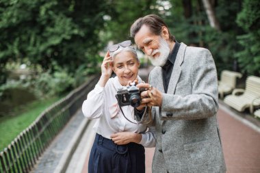 Yaşlı bir çift parkta dikiliyor ve kamerada fotoğrafları seyrediyor.