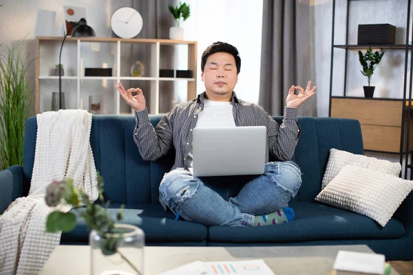 Asian man sitting on couch with laptop and meditating