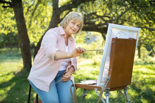 Happy Caucasian retired woman, wearing light shirt and denim jeans, painting on canvas and having fun at beautiful green garden or park on sunny day. Art, hobby and retirement concept