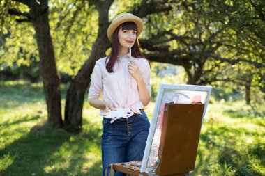 Yaratıcı resim, sanat, rahatlama ve ilham. Güzel, genç, bayan ressam gün batımında güzel, yeşil bir parkta veya bahçede resim yapıyor. Doğada açık hava atölyesi