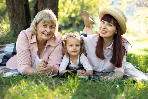 Grandmother, mother and grand daughter enjoying sunny garden holiday together outdoors, lying on green grass on blanket and smiling to camera. Leisure family lifestyle, happiness and moments