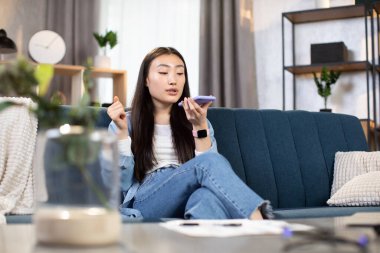 Genç Asyalı kadın telefonda hoparlörden konuşuyor, evdeki kanepede oturuyor.