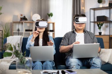 Kanepede oturan Asyalı çift VR kulaklıkları ve dizüstü bilgisayarlarıyla