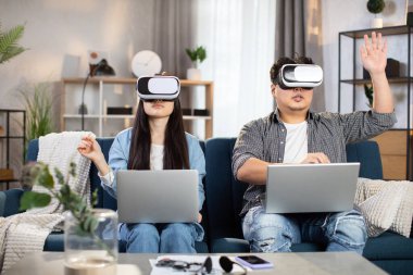 Kanepede oturan Asyalı çift VR kulaklıkları ve dizüstü bilgisayarlarıyla