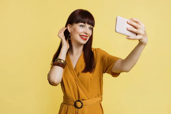 Excited in good spirits young woman, wearing trendy yellow suit, standing isolated on bright yellow colour background and doing selfie shot on mobile phone. People, technologies concept