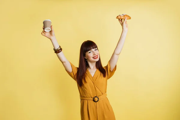 Good morning concept and lunch break. Happy excited jolly young woman in trendy yellow suit, having fun and holding coffee and croissant in hands raised, standing over yellow background Royalty Free Stock Images