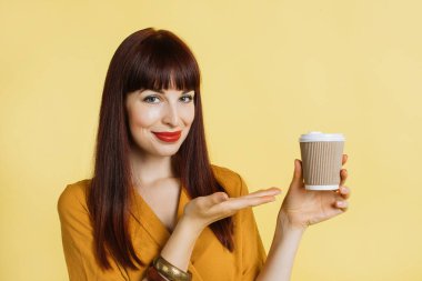 Parlak sarı gömlekli, kırmızı rujlu, gülümseyen kızıl saçlı beyaz kadın kahve fincanını alıp götürdüğünü gösteriyor. Sarı stüdyo arka planındaki neşeli genç kadın, boşluğu kopyala