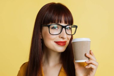 İnsanlar, duygular ve sıcak içecekler. Güzel gülümseyen, beyaz tenli, gözlüklü iş kadınının yüzünü kapatın. Sarı arka planda parlak kırmızı ruj ve kahve duruşu.