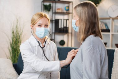 Profesyonel deneyimli hoş bayan doktor, yüz maskesi takıyor, yaşlı kadın hastasını muayene ediyor ve ev muayenesi sırasında steteskopunu kullanıyor.