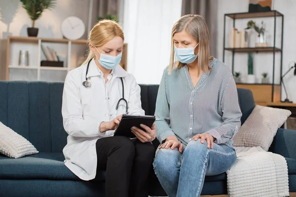 Doctor wearing safety protective mask consulting senior patient during home visit during covid-19 pandemic and flu outbreak. Doctor looking at the results and medications on tablet with senior patient