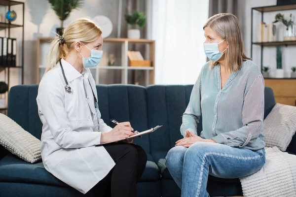 Female professional doctor in mask, making notes in clipboard, during homecare medical visit and talking with her senior woman patient in mask, sitting on sofa. Elderly people healthcare concept. Stock Photo