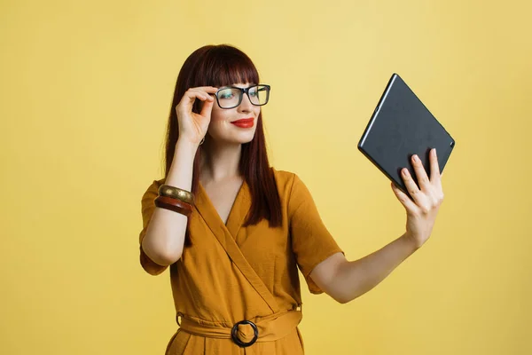 Jovem deslumbrante senhora de negócios bonita ou freelancer, vestindo roupas brilhantes da moda e óculos, segura tablet nas mãos, focado na leitura de notícias, relatório ou smth importante, fundo cor amarela — Fotografia de Stock