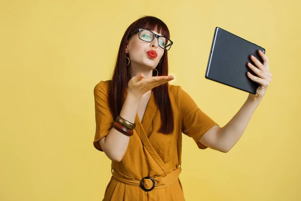 Pessoas, gadgets e emoções conceito. Linda menina bonita em óculos e vestido amarelo, enviando beijo de ar durante a chamada de vídeo, usando tablet digital no fundo do estúdio amarelo. — Fotografia de Stock