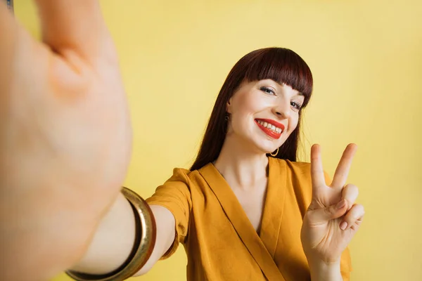 Sorrindo menina bonita de cabelos castanhos em bom humor, com maquiagem brilhante, batom vermelho, vestindo camisa amarela e pulseiras, posando fazendo selfie no fundo amarelo brilhante. — Fotografia de Stock