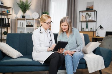 Genç kadın doktor yaşlı bir hastayı evde ziyaret ediyor, sağlık belgelerinin sigortasını anlatıyor, tedavi yöntemlerini tartışıyor, tabletteki ilaçların düzenlenmesini tartışıyor.