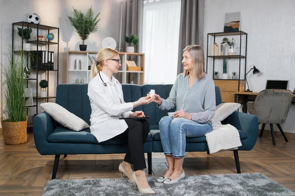 Healthcare, elderly patients, geratric deseases concept. Likable confident blond female doctor giving bottle with pills to senior female patient and telling how to take them during home visit