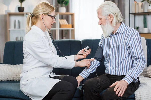 老年护理中的高血压。舒适的40岁金发女医生在家中用体能计测量老年男性70岁病人血压的画像. — 图库照片