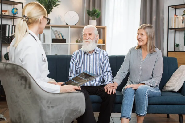 年轻的高技能女保健工作者，举行CT X光扫描，在家访期间咨询快乐的老年夫妇。退休夫妇坐在沙发上听医生讲话 — 图库照片