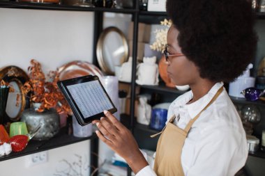 Afro-Amerikalı kadın elinde tabletle dükkanda envanter sayımı yapıyor.