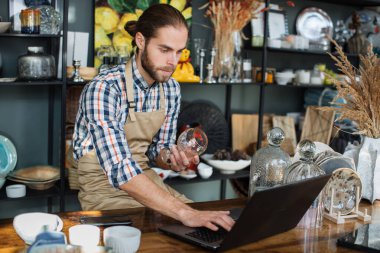Önlüklü sakallı adam dekor dükkanında dizüstü bilgisayarla çalışıyor.