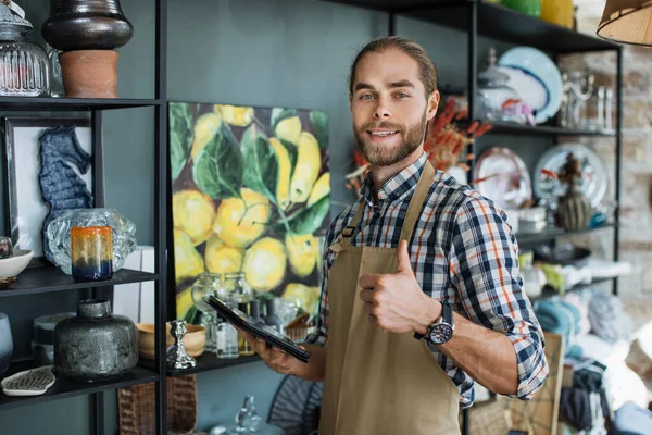 Vendedor sonriente mostrando el pulgar hacia arriba y sosteniendo la tableta en la tienda —  Fotos de Stock