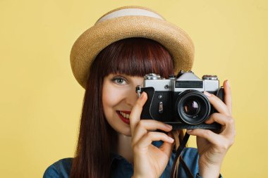 Hasır şapkalı güzel bir genç kadın fotoğrafçının fotoğraf çekmesini ve arka planda izole edilmiş sarı bir stüdyoda poz vererek retro dijital aynasız fotoğraf makinesine bakmasını.