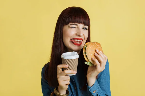 Nahaufnahme einer fröhlichen jungen attraktiven Frau, die in die Kamera grinst, Zunge zeigt, leckeren Burger in der Hand hält und Kaffee mitnimmt und ihre Mittagspause genießt. Isoliert auf gelbem Studiohintergrund. — Stockfoto
