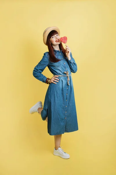 Fashion pretty girl in trendy jeans denim dress, white sneakers and straw hat, posing with one leg raised and big red lollipop, on yellow background