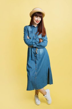 Kameraya poz veren kendine güvenen kız, sarı arka planda izole edilmiş. Kot pantolon ve hasır şapkalı, kollarını kavuşturarak poz veren şık bir kadının stüdyo fotoğrafı. Tam boy portre