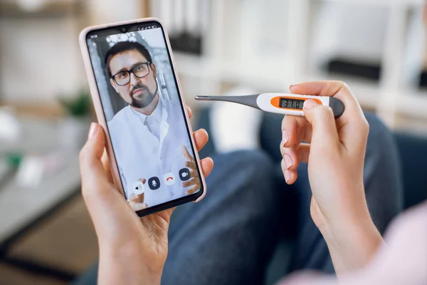 Online medicine concept. Close up screen view of confident bearded doctor communicates with his patient via a video call on a smartphone. Sick woman with thermometer takes gp remote consultation