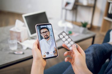 Yakışıklı, kendine güvenen bir erkek doktorla telefon ekranını kapat ve hastaya video görüşmesiyle uzak bir konsültasyon yap. İnternette danışmanlık için kadın eller hap ve cep telefonu tutuyor.