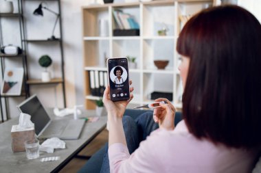 Hasta kız ve tıbbi internet danışmanı. Bir kadının akıllı telefonuyla doktorunu ararken, evde otururken, ateşi yüzünden kötü hissederken, elinde termometreyle koltukta otururken..