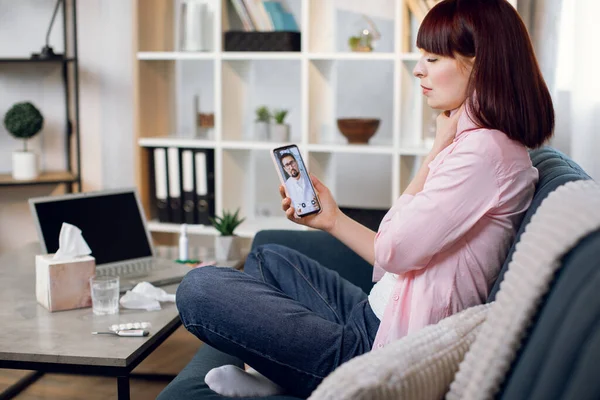 Telemedicine, online appointment concept. Side view of pretty young sick woman, sitting on sofa at home, suffering from throat pain, having video chat with male doctor by phone