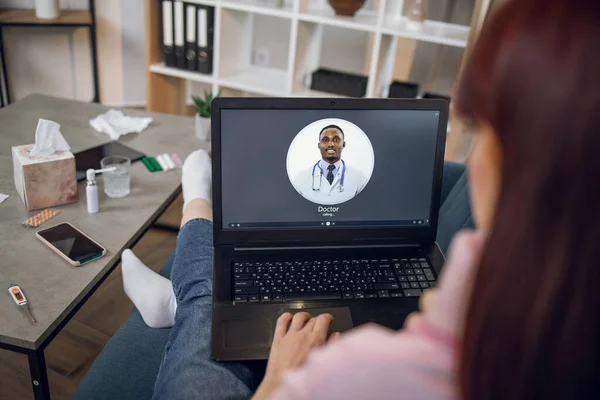 Over shoulder videocall view. Close-up of laptop screen with medical application for remote service. Unrecognizable female patient calling to male black doctor using laptop at home