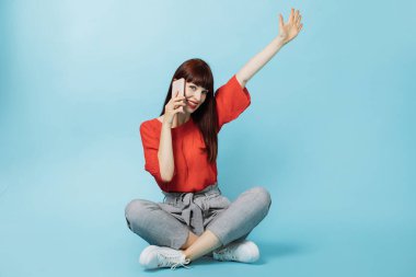 Uzun düz kızıl saçlı, mutlu bir kadın, parlak resmi kıyafetler giymiş, Lotus pozu vermiş, tek kolu havada, mavi arka planda, konuşan bir cep telefonu.