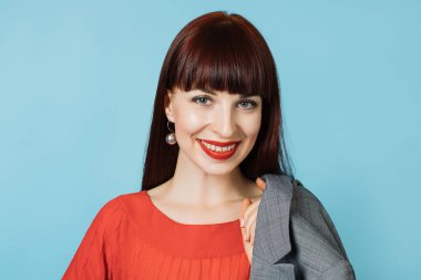 Kırmızı tişörtlü, parlak kırmızı rujlu, omuzlarında gri ceketi olan, izole edilmiş stüdyo mavisi arka plandaki kameraya gülümseyen genç ve çekici kızıl saçlı kadının portresini yakından çek.