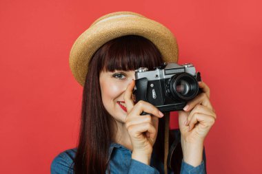 Hasır şapkalı güzel bir genç kadın fotoğrafçının fotoğraf çekmesini ve arka planda izole edilmiş kırmızı arkaplanda poz vererek retro dijital aynasız fotoğraf makinesine bakmasını.