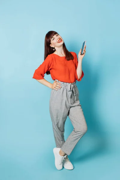 Full length portrait of young likable pretty woman in fashionable formal wear, gray pants and red shirt, holding cell phone in hands, posing on isolated blue background
