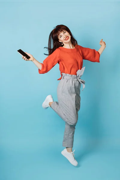 Retrato de uma mulher de cabelos vermelhos salto engraçado na moda roupas brilhantes, segurando smartphone na mão, se divertindo isolado sobre estúdio fundo azul — Fotografia de Stock