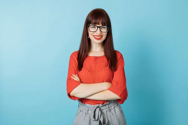 Foto de jovem empresária bastante confiante gerente em blusa vermelha brilhante e óculos, posando para câmera com braços cruzados, fundo de cor azul isolado. Cintura para cima retrato — Fotografia de Stock