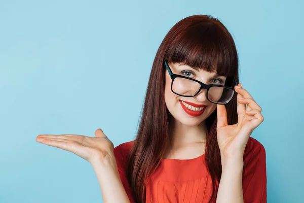 Retrato de mulher de negócios bonita em camisa vermelha e óculos, mostrando algo imaginário, espaço de cópia vazio para algum texto, produto ou slogan, na palma da mão aberta, isolado em fundo azul — Fotografia de Stock
