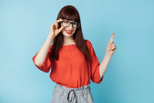 Jovem empresária em elegante blusa vermelha brilhante e óculos apontando para cima no espaço livre cópia, sorrindo para a câmera no fundo azul — Fotografia de Stock
