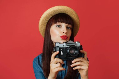 Hobi, fotoğraf çekme, seyahat konsepti. Şapkalı genç bir kadın, fotoğrafçı amatör, gülümseyen ve hava öpücüğü veren, renkli kırmızı stüdyo arka planında, eski model bir kamerayla fotoğraf çeken..