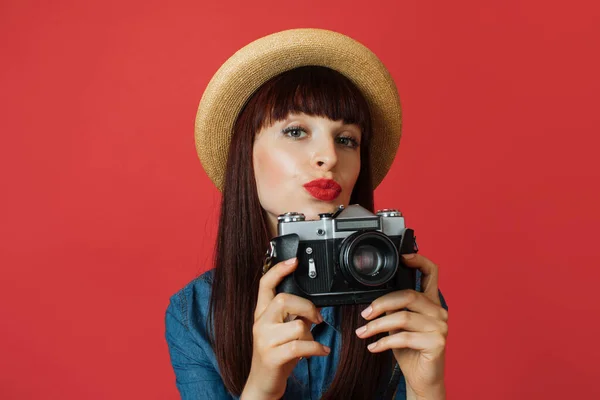 Hobby, taking photos, travel concept. Gorgeous young woman in hat, photographer amateur, smiling and making air kiss, taking picture on old vintage camera, on colorful red studio background.