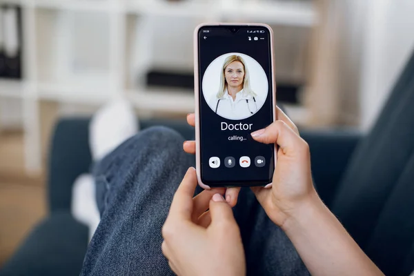 Virtual medical consultation. Unrecognizable woman, sitting on couch at home, calling to doctor via video call on cellphone. Close up of smartphone screen with picture of blond mature female doctor.